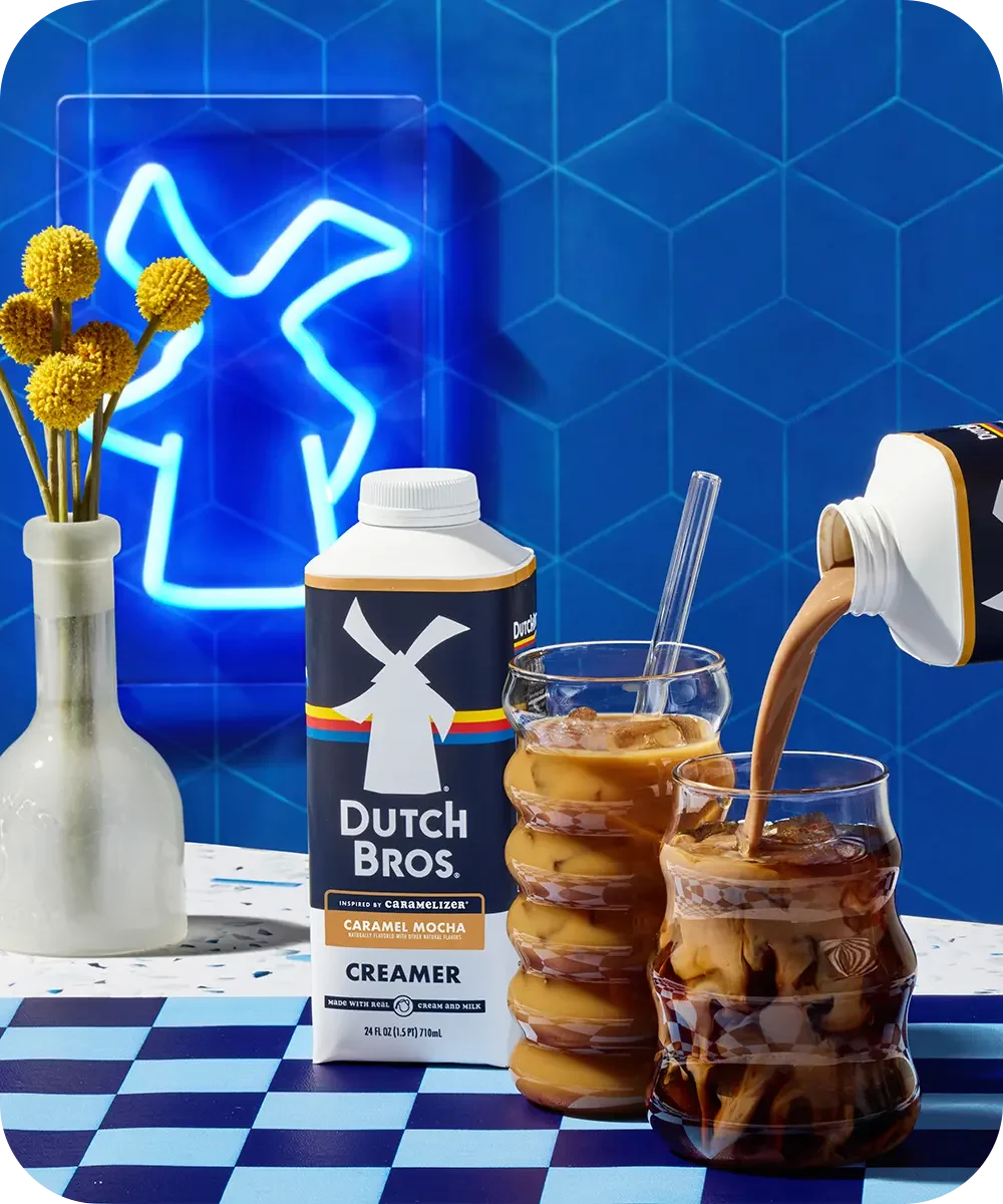 Dutch Bros Caramel Mocha creamer pouring into iced coffee against blue tile wall with neon sign and yellow flowers.