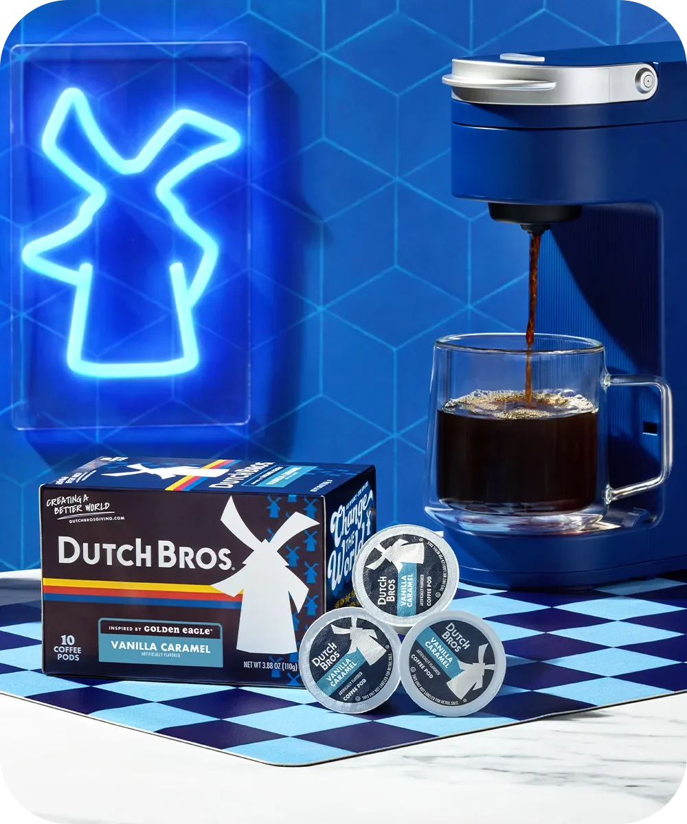 Blue coffee maker brewing into glass cup with Dutch Bros coffee pods and box against blue windmill neon sign backdrop.