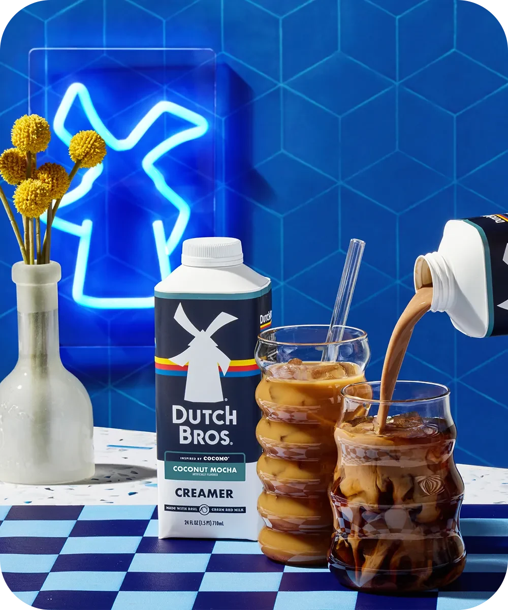 Dutch Bros creamer pouring into iced coffee drinks with blue neon sign and yellow flowers against geometric blue tiles.