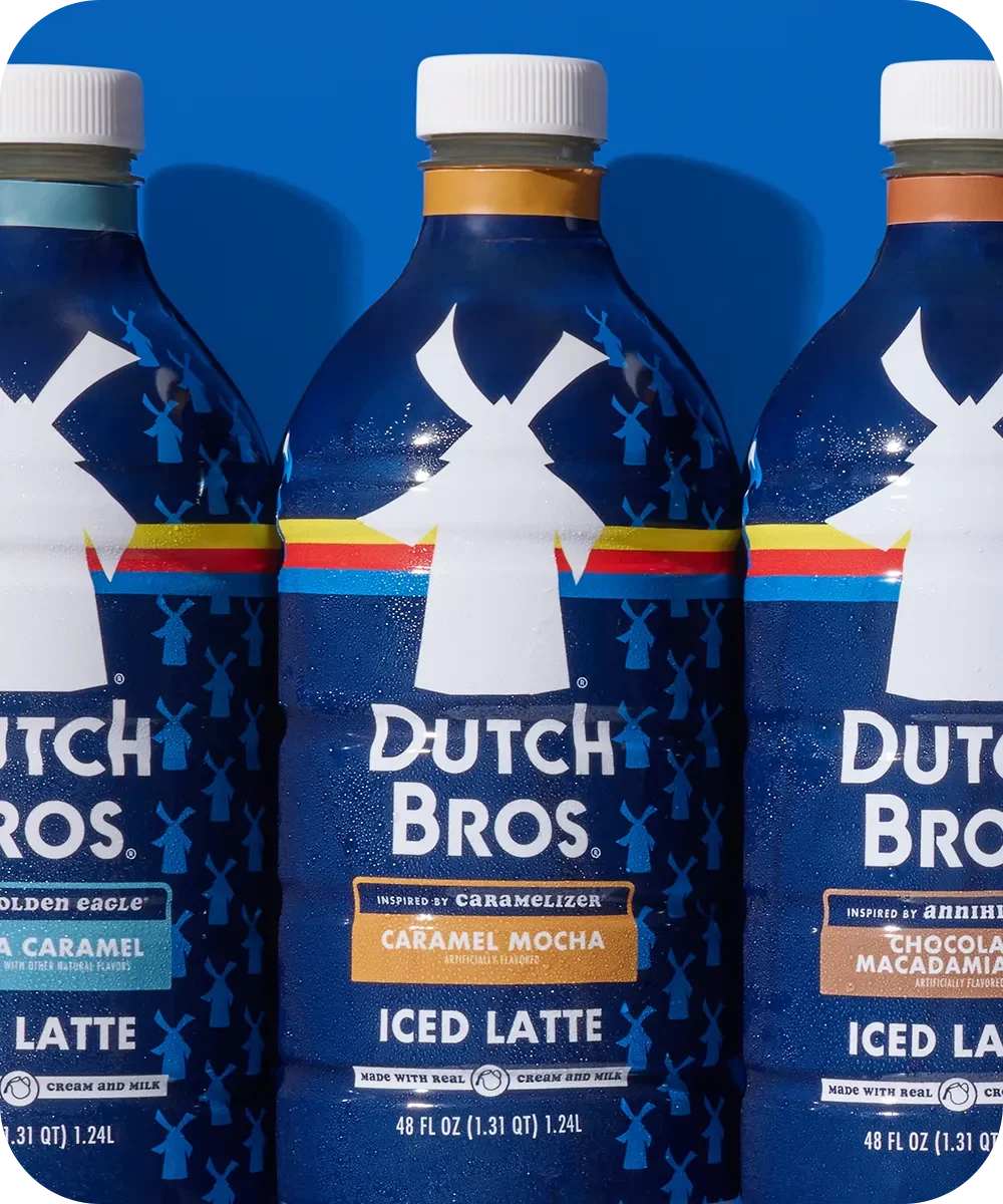 Three Dutch Bros bottled iced lattes in dark blue packaging with different flavors against a blue background.