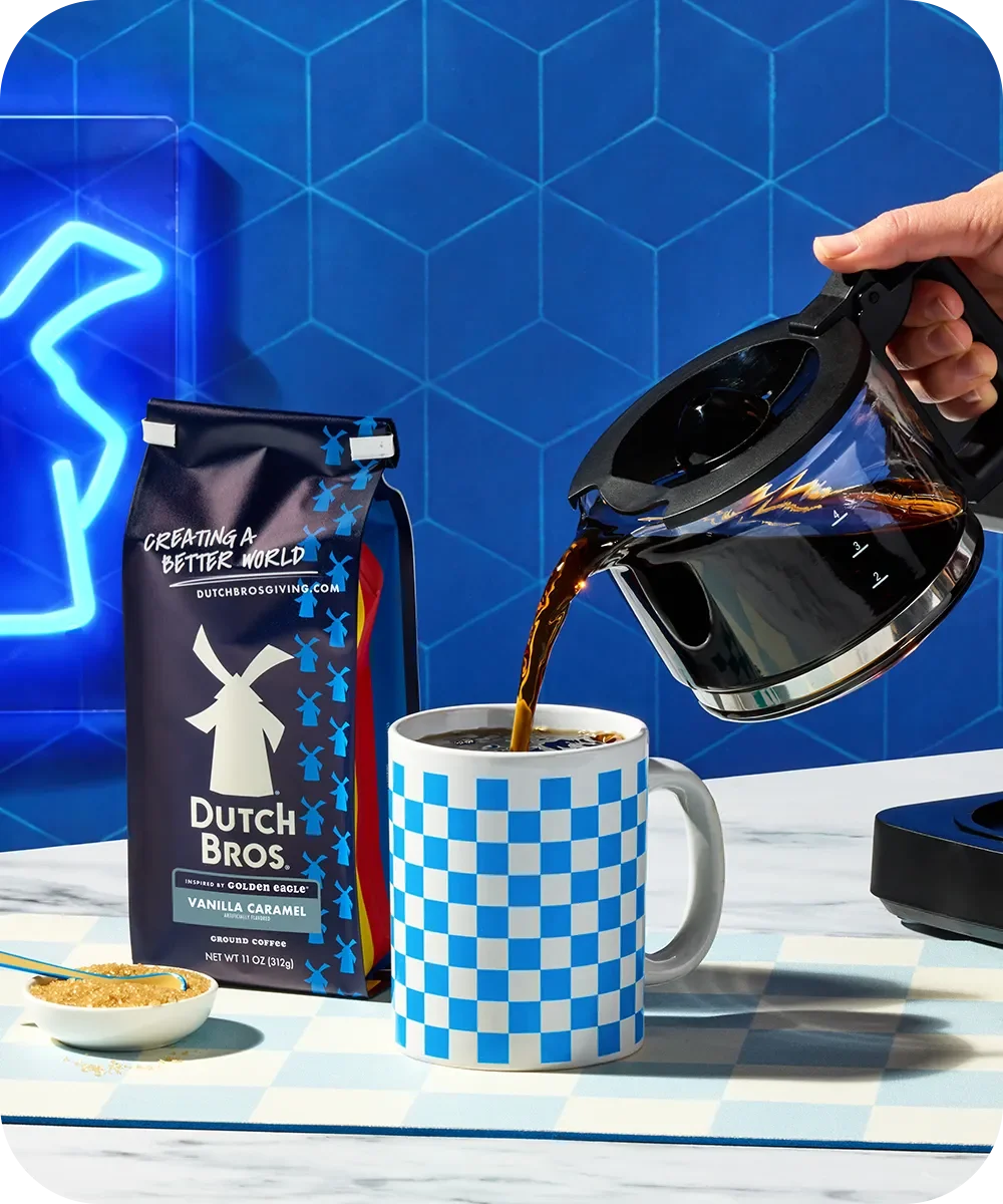 Hand pouring coffee from a pot into a blue checkered mug next to Dutch Bros coffee bag against blue geometric background.