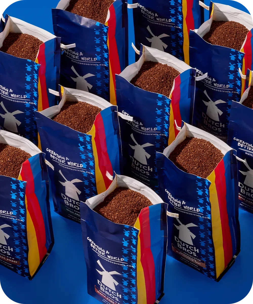 Blue packages of Dutch Bros coffee with colorful stripes and windmill logo, filled with ground coffee beans.