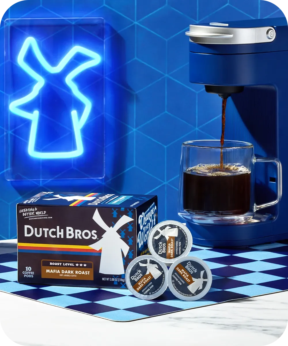 Blue coffee maker brewing Dutch Bros dark roast coffee with windmill logo neon sign and coffee pods on checkered surface.