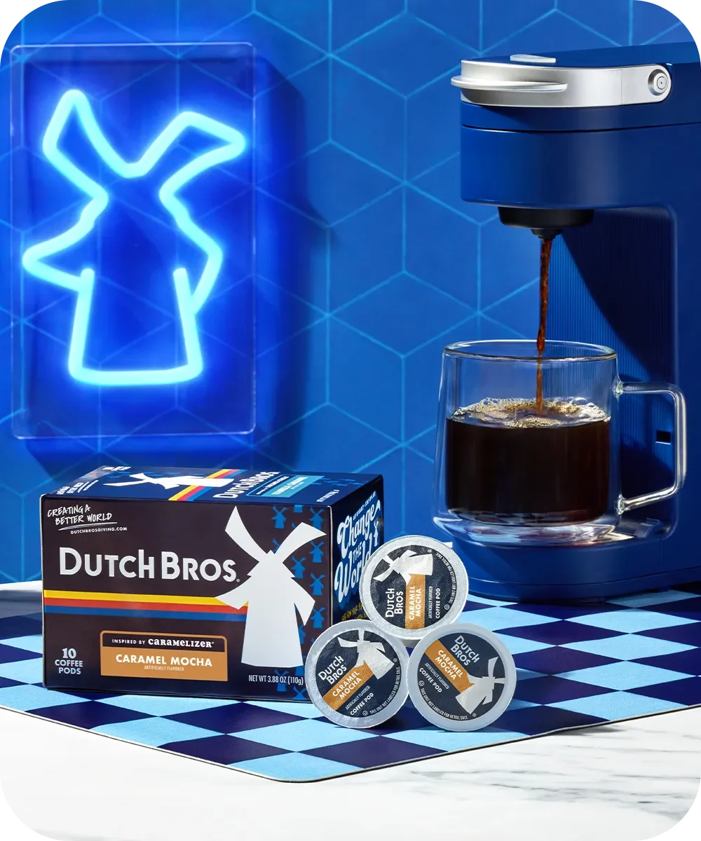 Coffee machine brewing Dutch Bros Caramel Mocha with coffee pods and packaging against blue windmill logo backdrop.